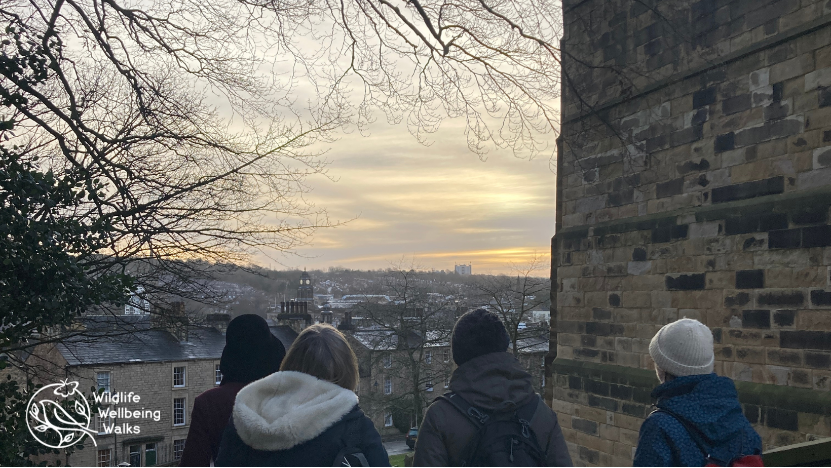 4 people looking towards a sunrise with a castle wall and overhead branches of a leafless tree in silhouette.
